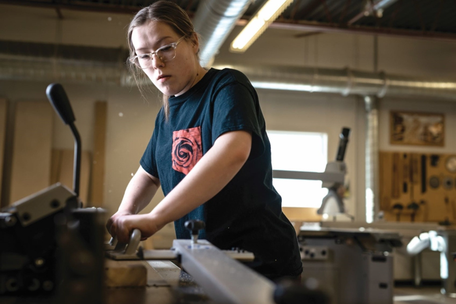A person wearing a black t-shirt pushes wood through a saw in a woodshop